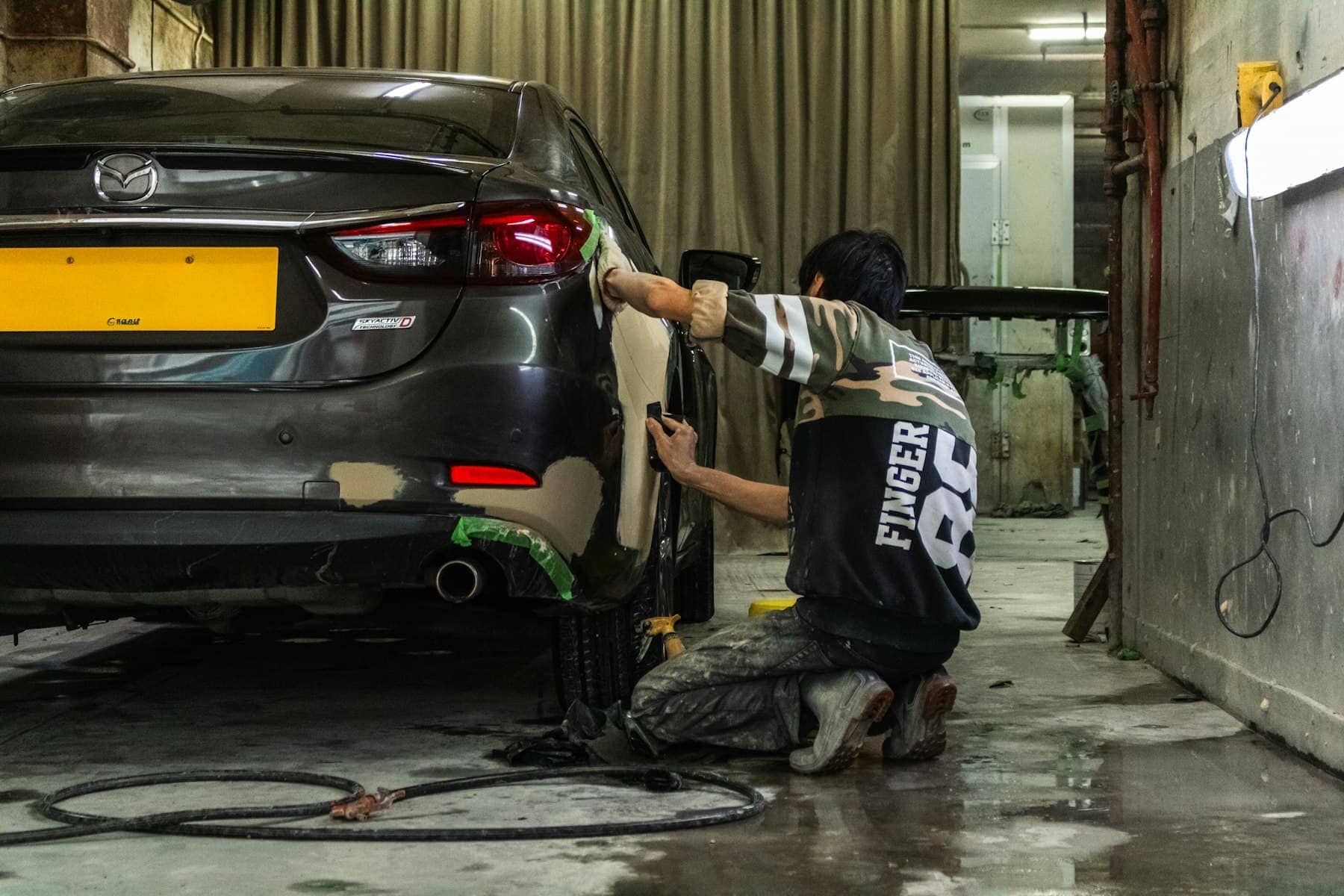 Technician sanding the rear panel of a grey sedan in a body shop