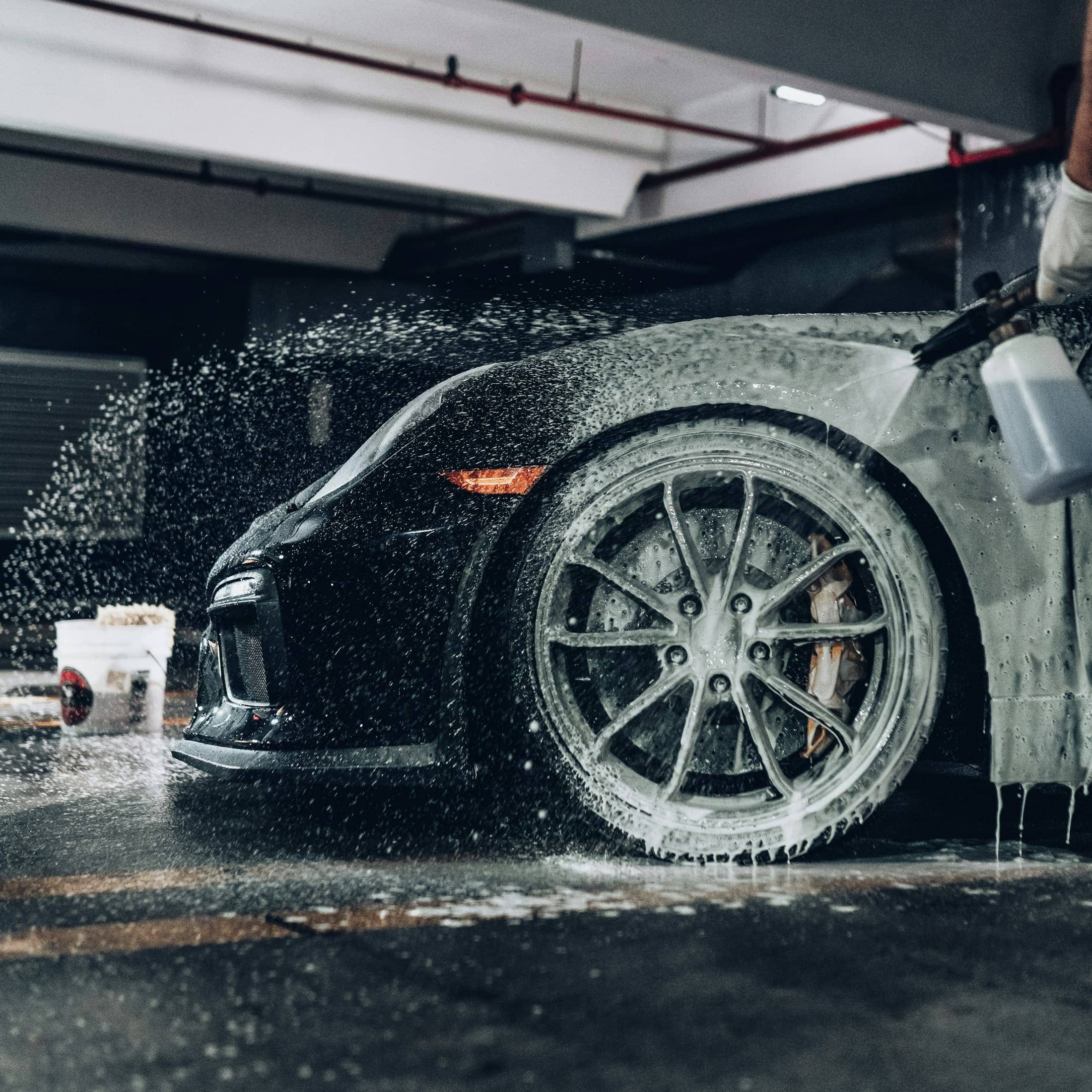Dark sports car being sprayed with snow foam during a wash in a covered garage