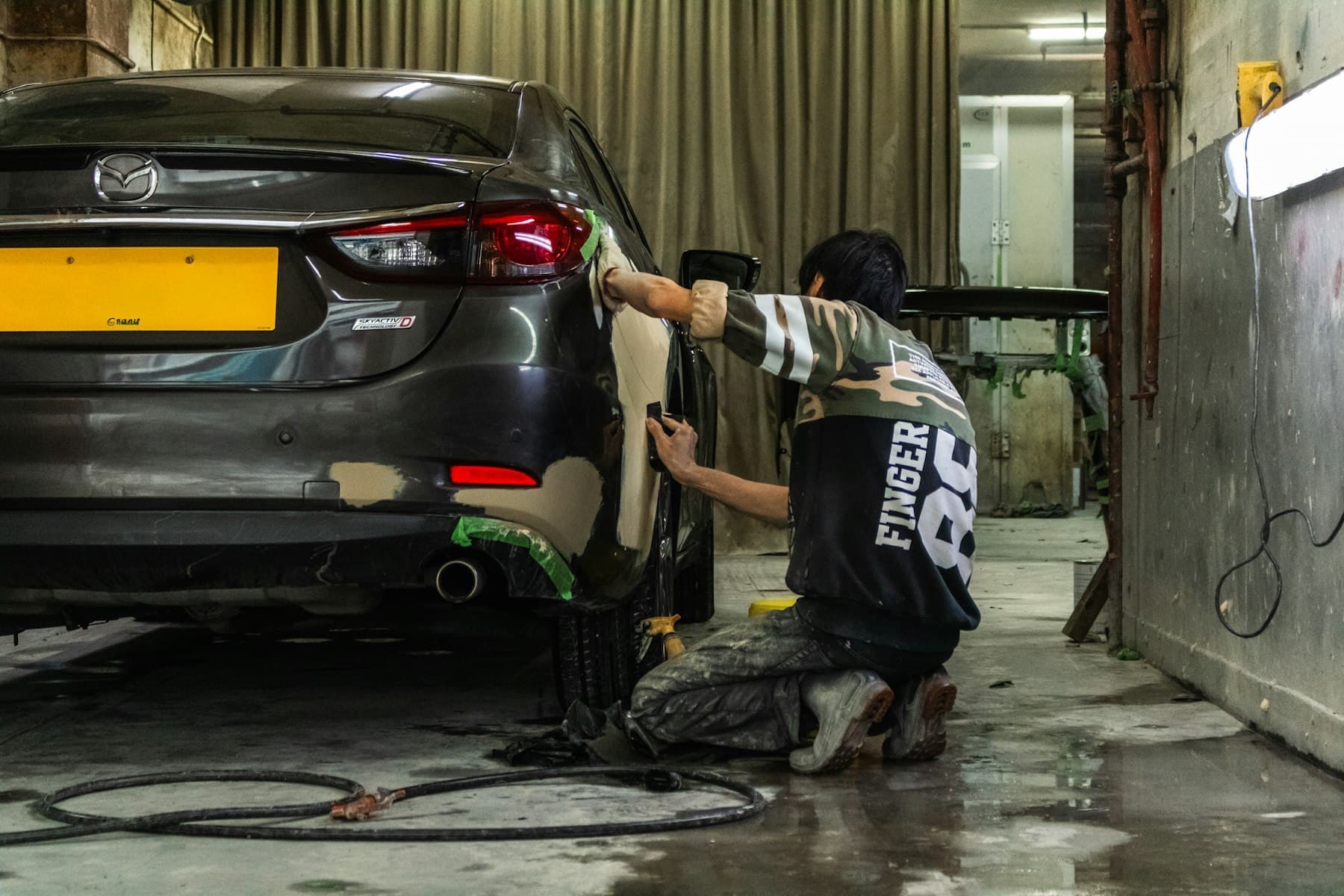 Technician sanding the rear panel of a grey sedan in a body shop